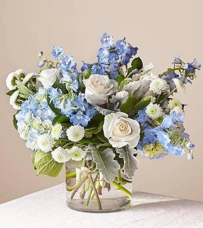 Clear Skies™ Bouquet by Flower Co. is a design in soft white and blue tones with premium roses, delphinum, dusty miller and chrysanthemum that transmits serenity and hope. 